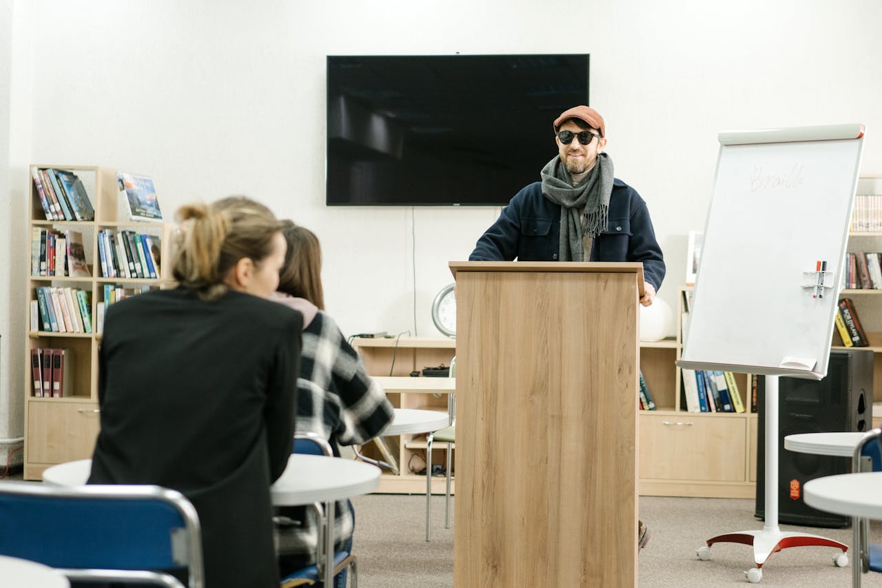 A man with sunglasses conducts a presentation on Braille in a library setting, engaging attentive listeners.